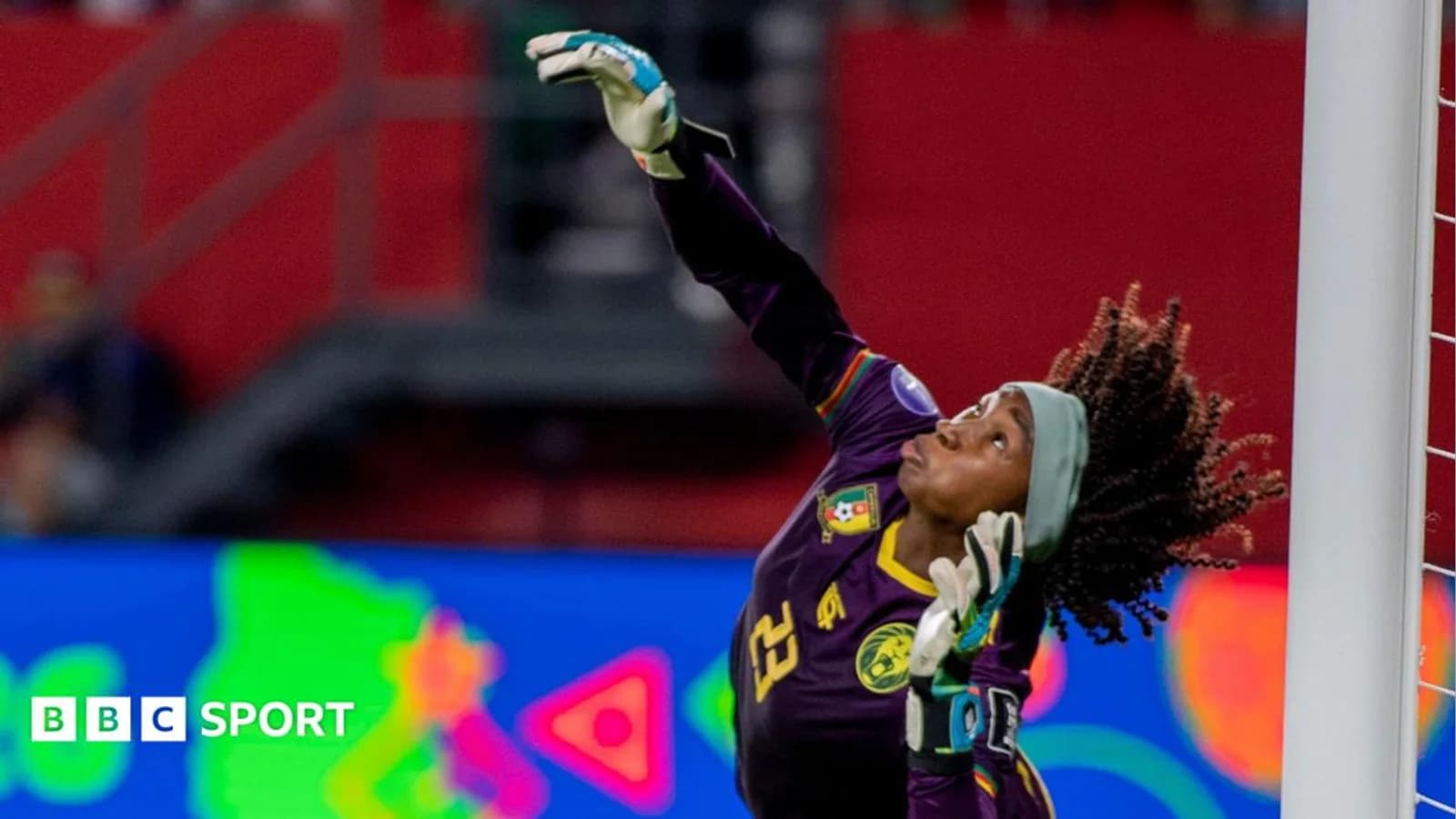 Cameroon goalkeeper Michaely Bihina, wearing an all-purple kit and wide blue headband, arches her back as she raises her right arm to push away a penalty · Fuente: BBC Sport Football
